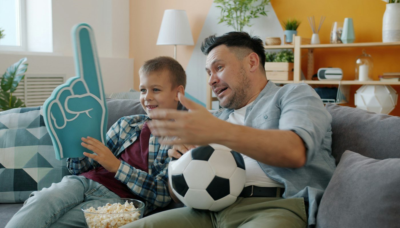 Vader en zoon op de bank met een voetbal, een schuimen vinger en popcorn, druk pratend over de wedstrijd