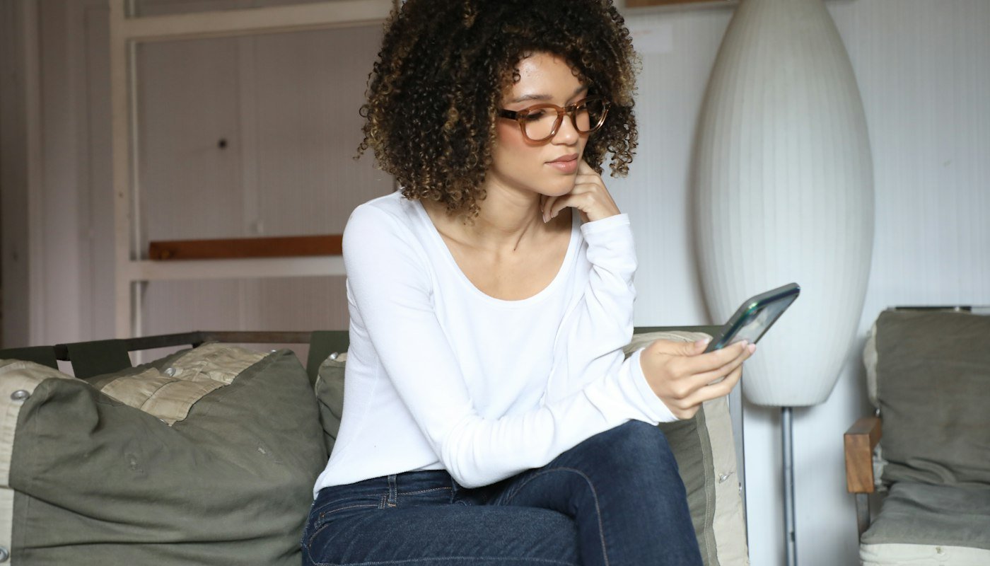 Een vrouw met een bril zit op de bank en kijkt op haar smartphone