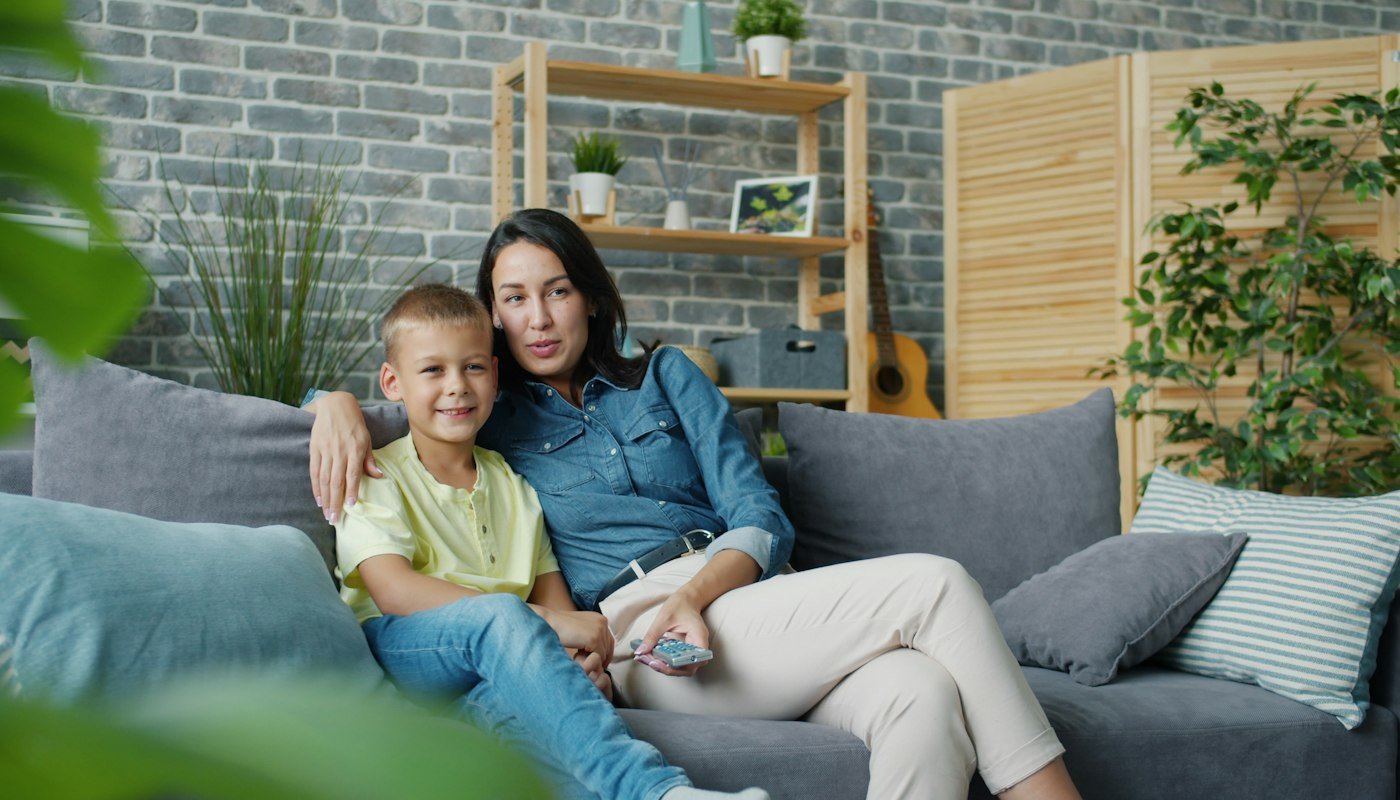 Moeder en zoon samen op een grijze bank in een woonkamer met planten en een boekenkast