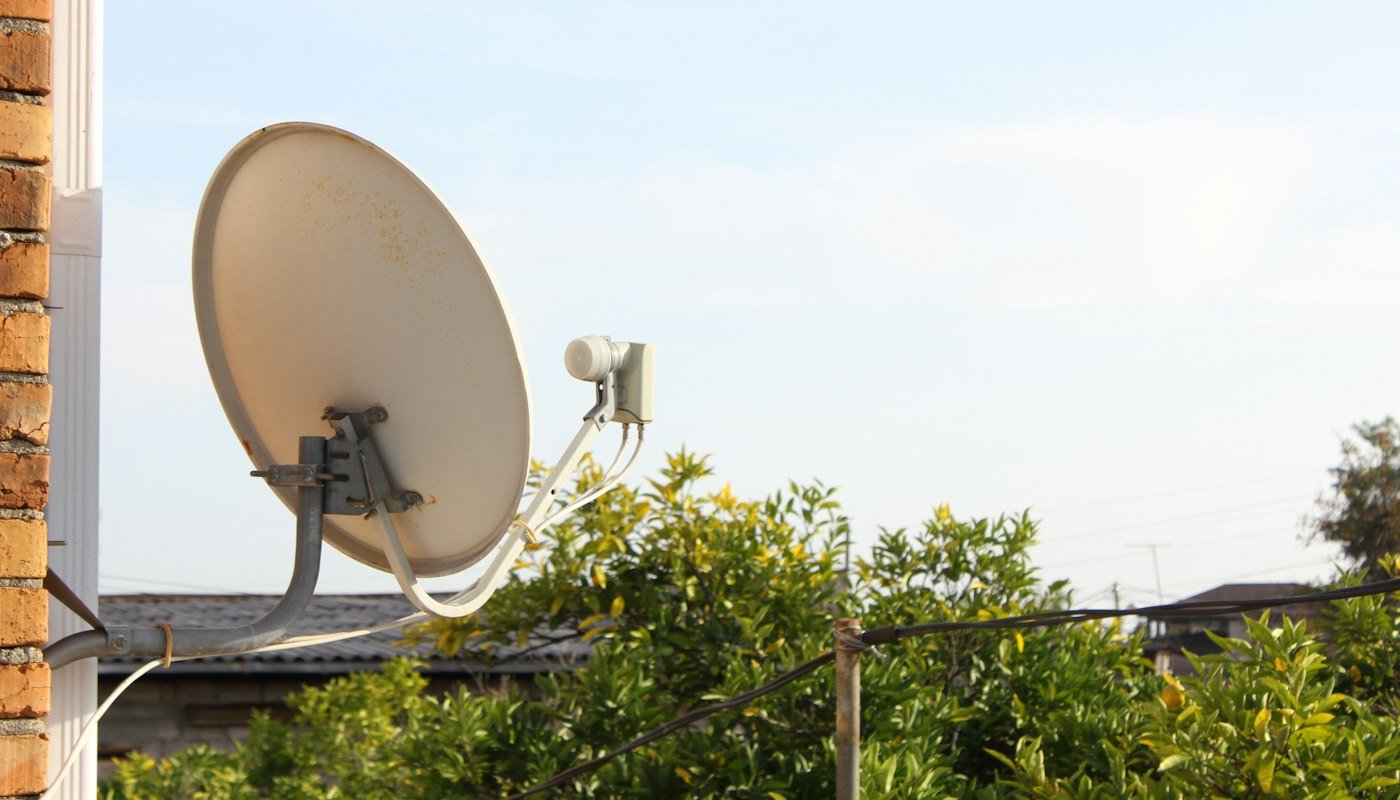 Een satellietschotel met LNB aan een bakstenen muur, met bomen en daken op de achtergrond